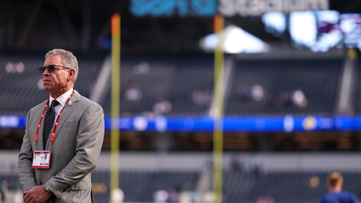 Troy Aikman em um estádio de futebol americano; sua relação com os Dolphins é tema de análise sobre o futuro na televisão.